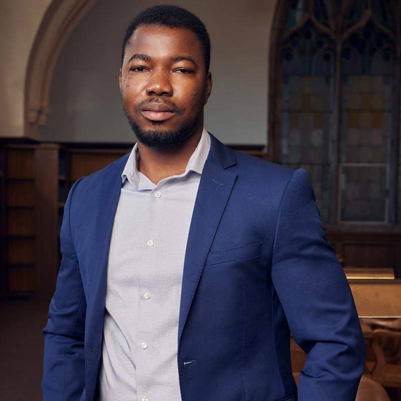 Joseph Kangmennaang stands in a library with a stained-glass window behind him.