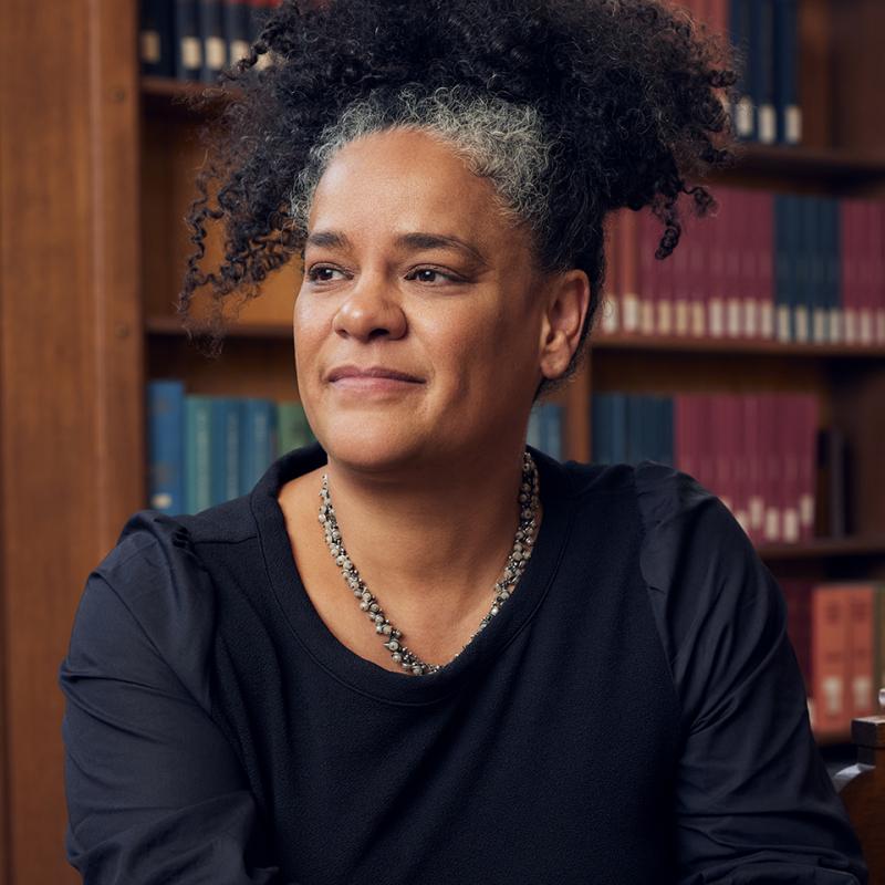 Katherine McKittrick sits at a library table with a wall of books behind her. Her hands are folded in front of her and rest on the table.