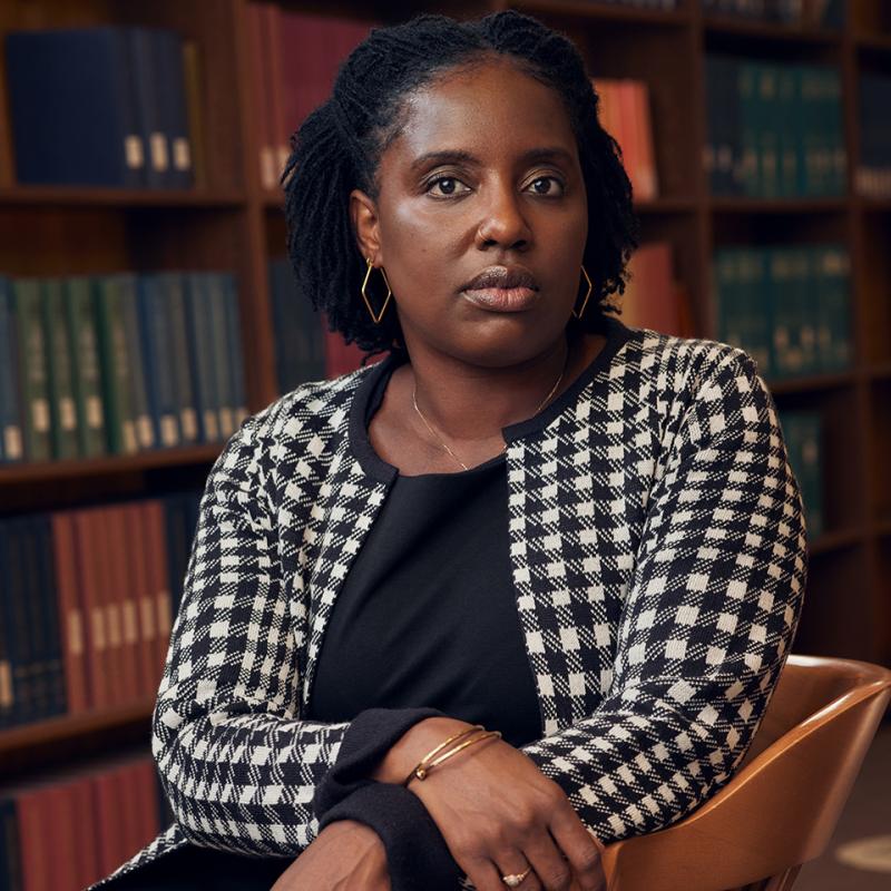 Kesha Fevrier sits in a library chair with a wall of books behind her. Her arms cross over her front and rest on the art of the chair.