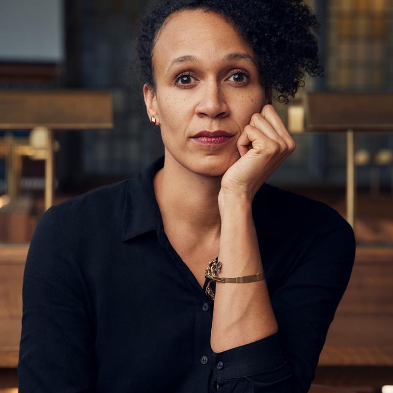 Vanessa Thompson sits at a library table. Her right arm is across her lap and her left elbow rests on the back of a chair while her hand is fisted alongside her chin.