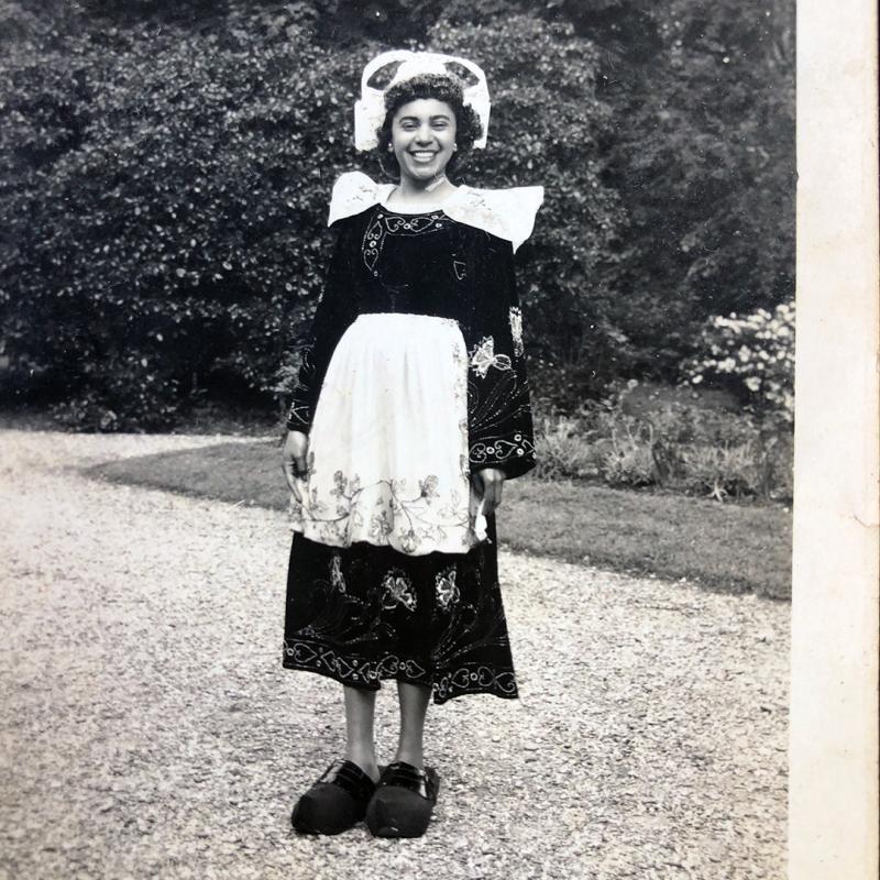 An old stained, black and white photo of Elizabeth Kawaley wearing an embroidered dress, apron, and hat. She is wearing clogs and standing in front of some trees.