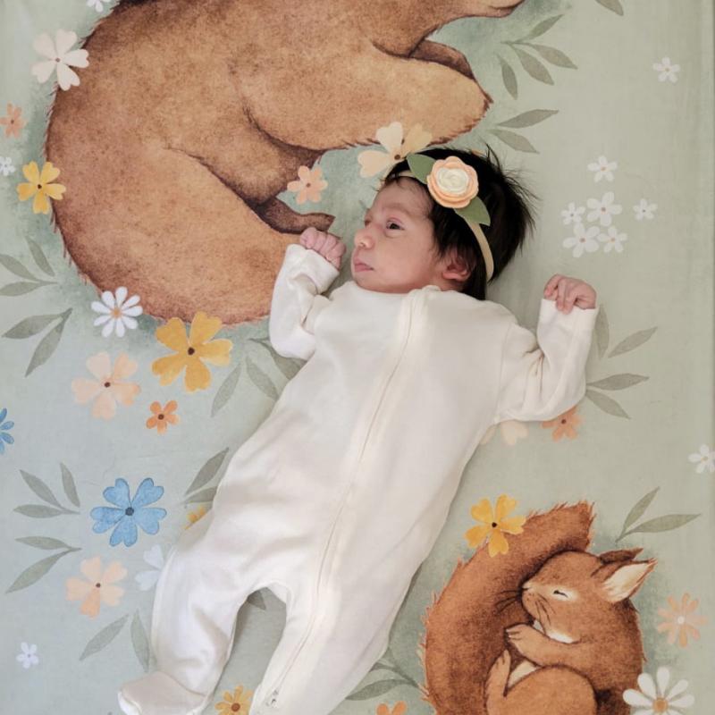 A baby, with full head of hair, lies on a blanket printed with forest animals.