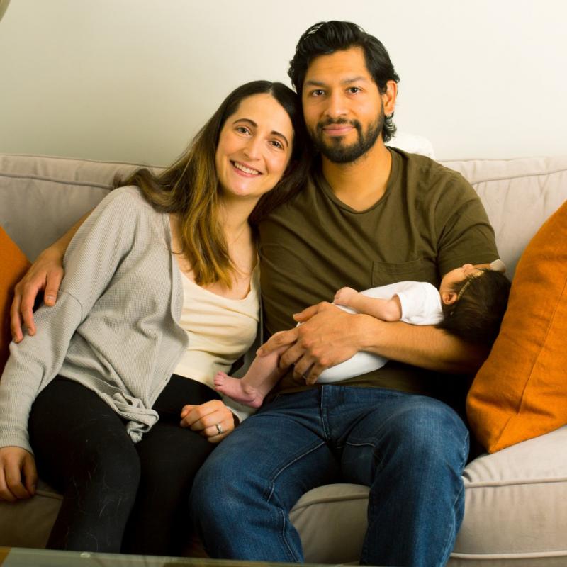A couple sits on a couch with the man cradling a newborn baby.