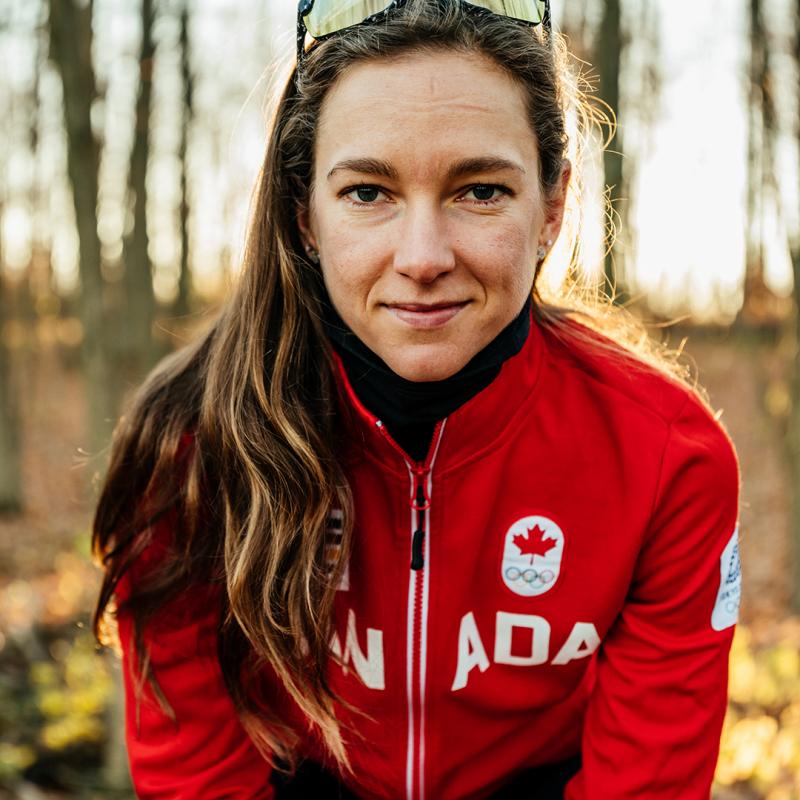 Portrait of a woman taken in a wooded area. She seated andis looking at the camera and wearing an olympic jacket and sunglasses on top of her head.