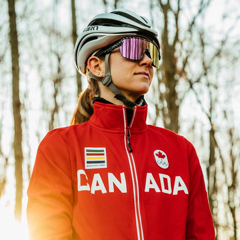 Woman standing in a wooded area with the sun setting behind her. She is wearing an olympic jacket, sunglasses, and a bike helmet. She is looking off to the side.