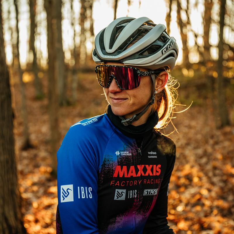 An upper body portrait of a woman sitting on her bicycle in a wooded area. She wears a biking shirt, sunglasses, and a bike helmet. She is looking to her right with the sun setting behind her.
