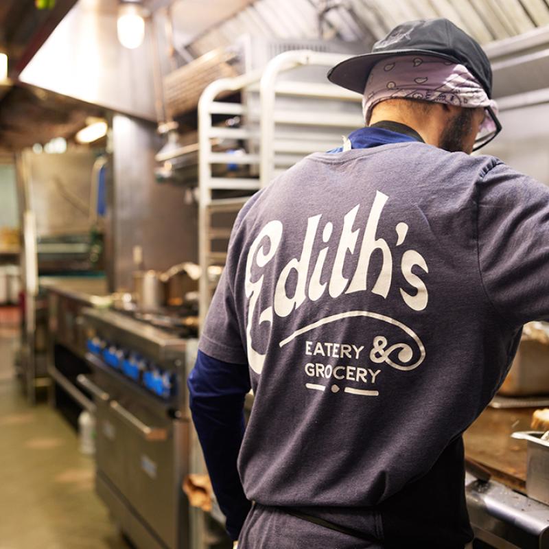 An Edith's deli employee stands in the kitchen with his back to the camera. He is wearing an Edith's Eatery and Grocery t-shirt.