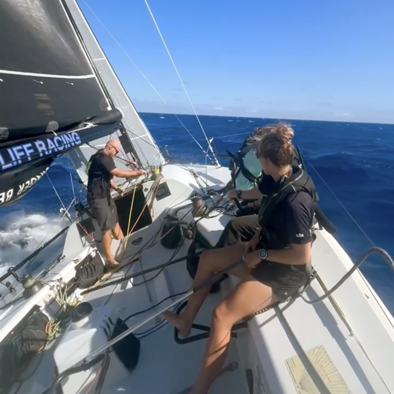 Members of the Oakcliff Black crew sit atop a sail boat. The boat is tilting to the left.