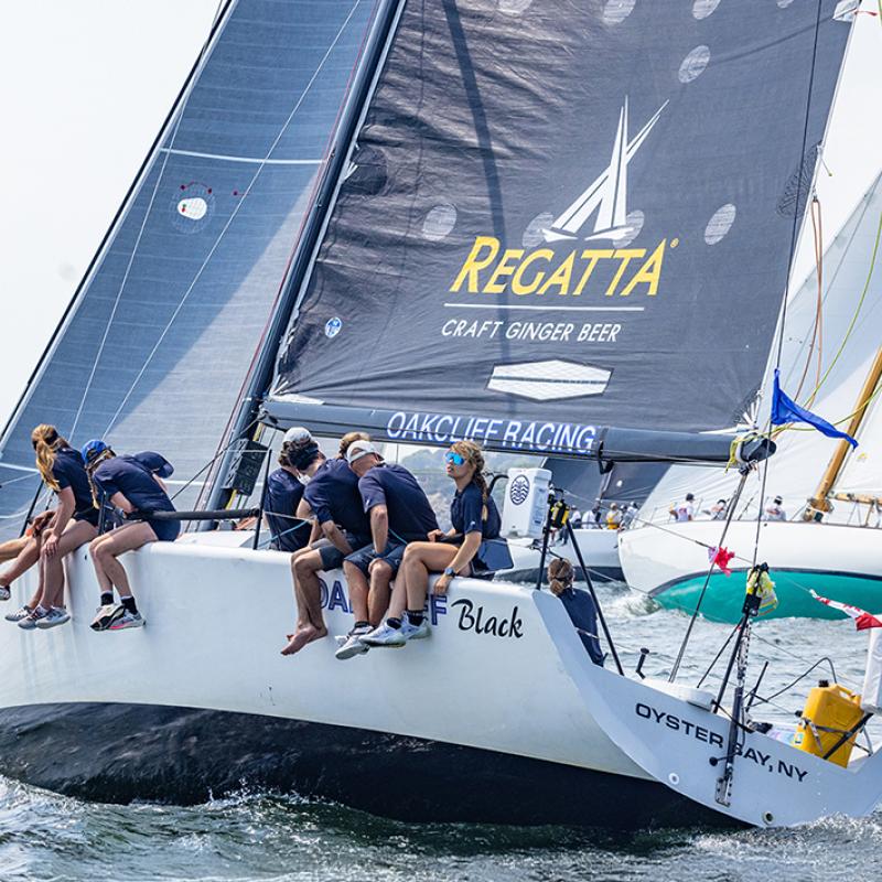 Members of the Oakcliff Black crew sit on the side of the boat, their legs are hanging off the left side. Behind them are other racing boats.