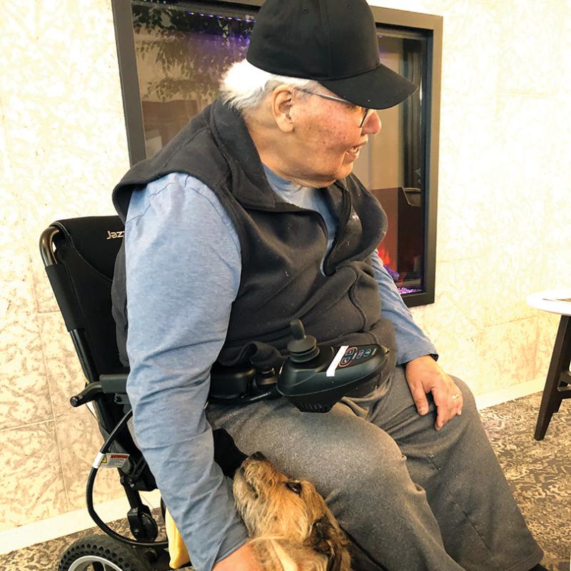 Murray Sinclair seated in a motorized wheelchair pets a small tan dog resting against his leg near a fireplace.