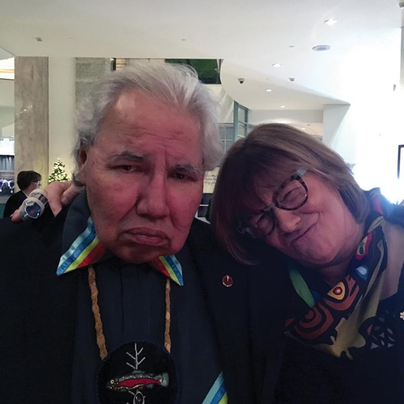 Murray Sinclair and Shelagh Rogers lean their heads together at an indoor event, both wearing formal attire and patterned scarves.