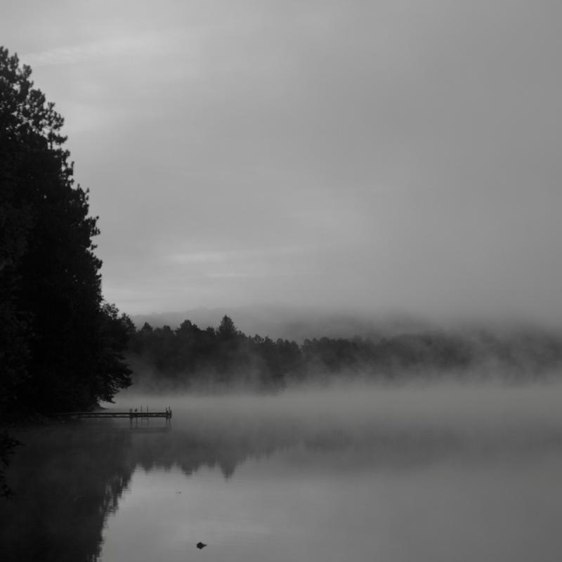 Black-and-white photograph of a fog-covered lake and treeline reflected in still water.