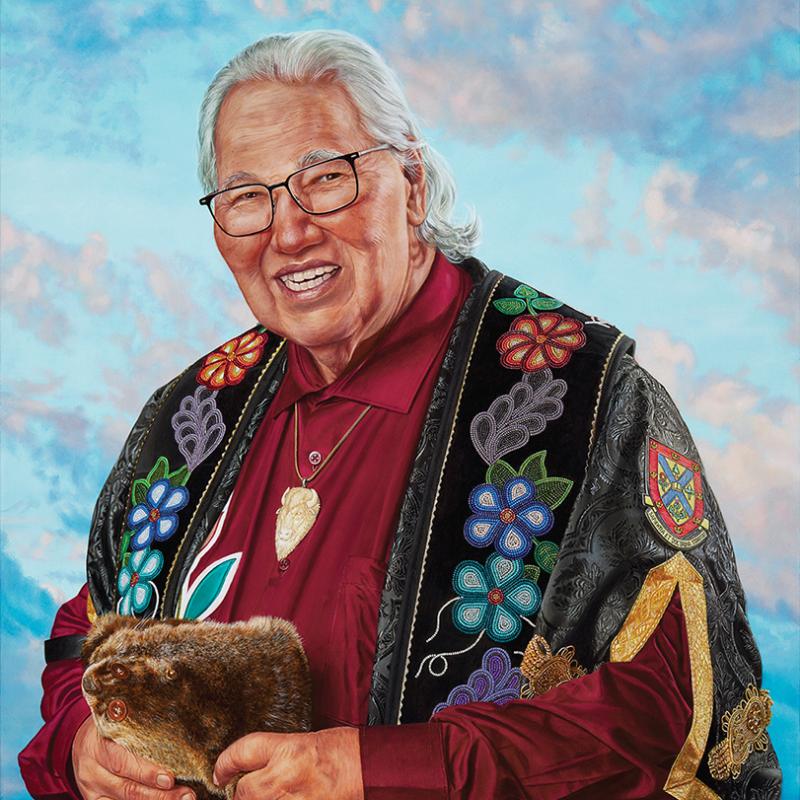 Portrait of a smiling Murray Sinclair wearing glasses and a colourful floral-beaded garment, holding fur against a soft blue and pink background.