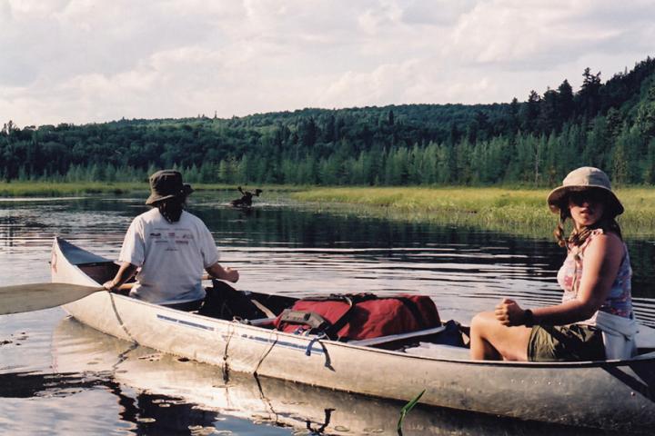 Two students paddle a canoe on a lake lined with evergreen trees.
