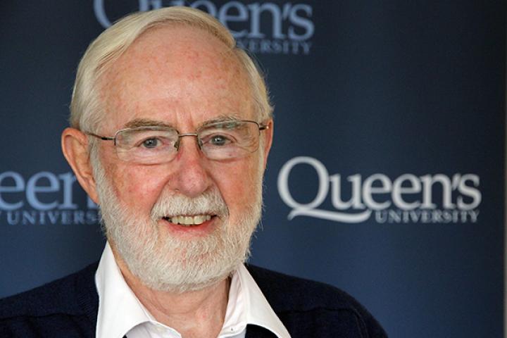 Photo of Arthur McDonald in front of a Queen's blue backdrop, looking at the camera and smiling.