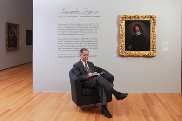 Carlos Prado reads a book while sitting in a chair. Descartes as painted by Nason hangs on a wall behind him.