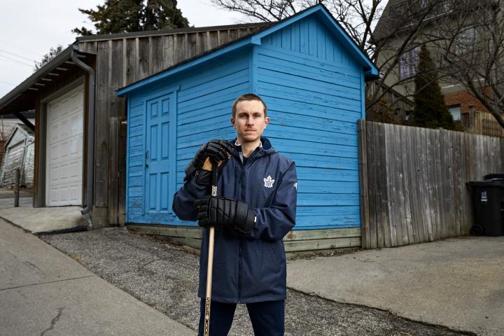 Darryl Metcalfe standing in front of a blue wooden shed holding a hockey stick and wearing hockey gloves.