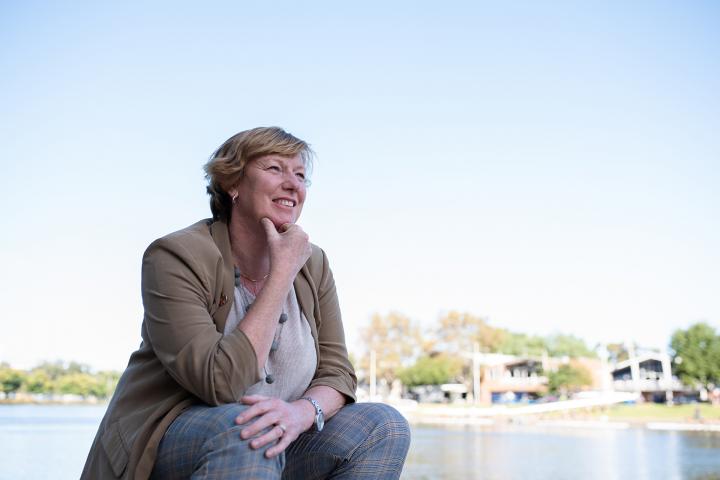 Dr. Alison Baker, wearing a blazer and plaid pants, sits by the water's edge with one hand laying across her legs and the other touching her chin.