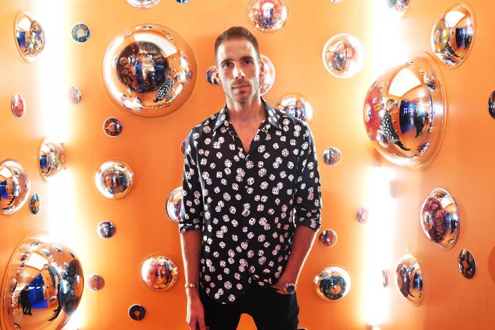 Simon Huck, in a black shirt with a dice pattern and black pants, standing in front of an orange backdrop covered in bubbles.