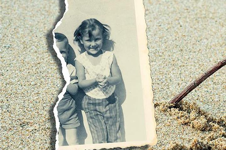 Book cover – torn black and white photo of two little girls, only the face of one girl is now visible, sitting on top of sand