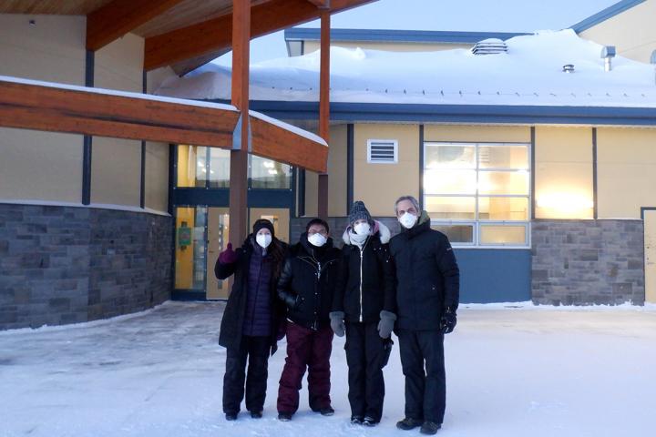 Four people are standing outside a building with winter coats, scarfs, gloves, and face masks on