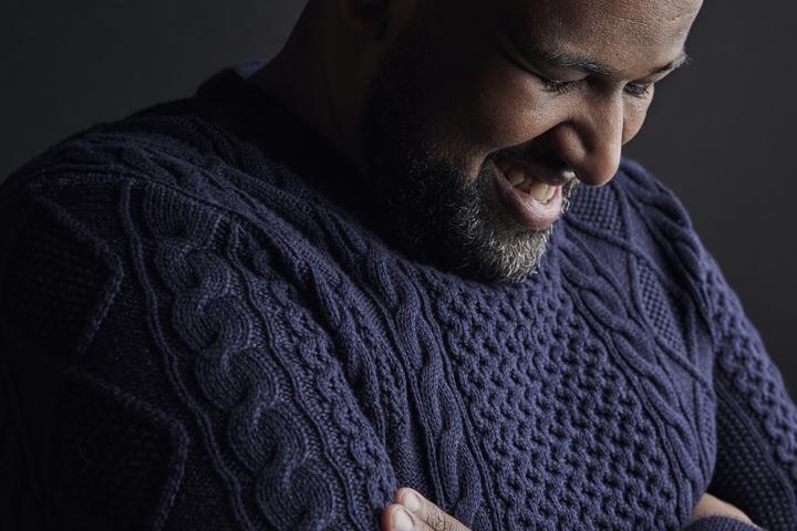 Elamin Abdelmahmoud, wearing a black knit sweater, stands against a black background, facing to his left, with his arms crossed, looking down and laughing. 