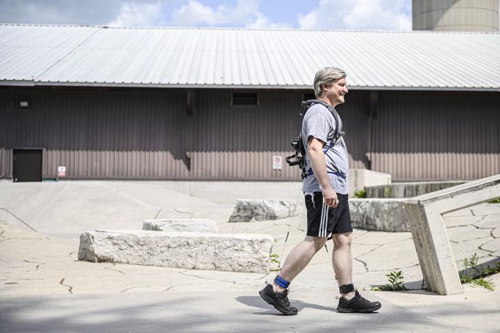 A man walks with the exoskeleton on his back and cables attaching to his legs.