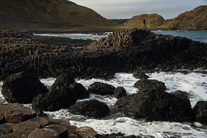A man stands on rocks in the ocean with green hills behind him
