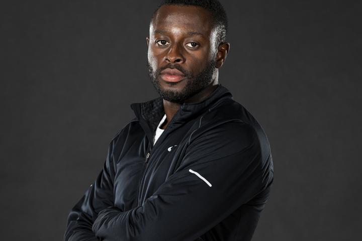 Sam Effah, arms crossed against his chest, holding a pair of running shoes, against a black backdrop.