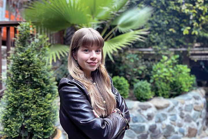 Young woman with long blond hair, wearing a leather jacket, stands in front of a garden.