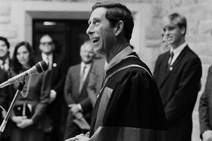 A man stands in front of a microphone speaking to a small crowd, wearing a graduation gown.