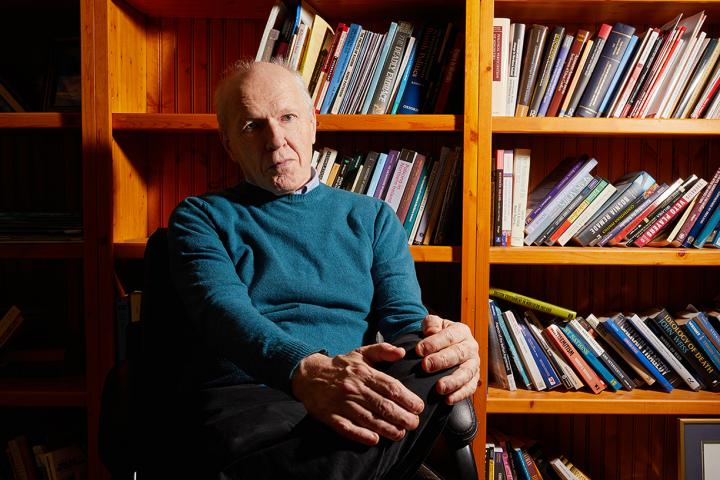 John McGarry sits in an office chair in front of a wall of bookcases. His legs are crosses with his hands resting on his left knee.
