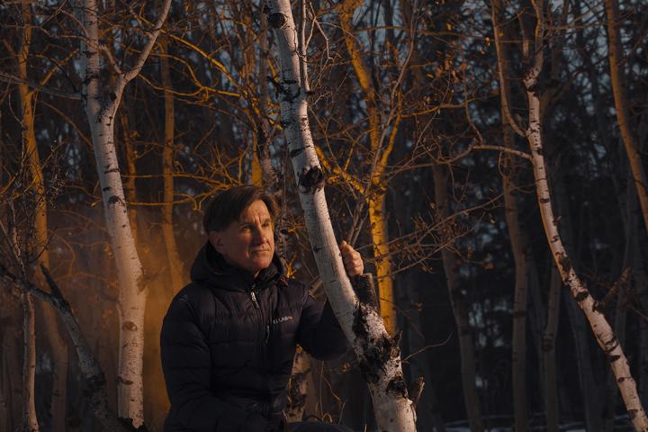 Edward Struzik is crouching in the woods wearing a winter jacket. He is surrounded by trees with a reflection of the light from a fire appearing behind him.