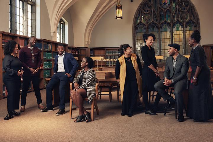 Eight members of the Black Studies faculty talk to each other in a group in a library. The library features a vaulted ceiling, arches, and stained glass windows.
