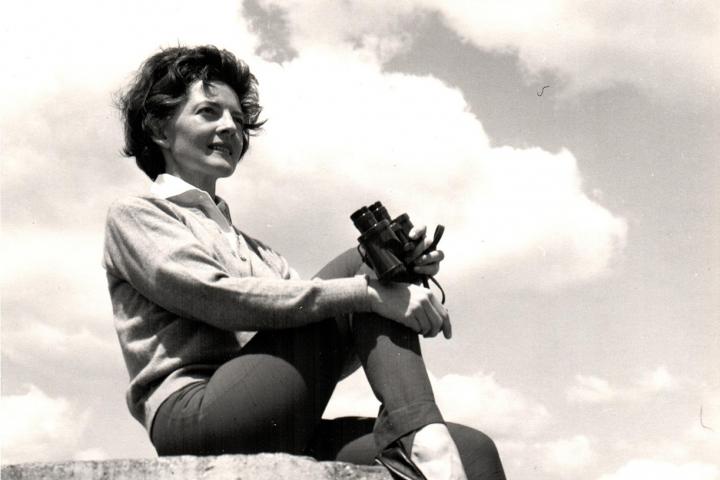 A photo from the 1960s shows a woman sitting on a large rock. Her right leg is pulled up towards her body and her hands are holding her knee. In her left hand is a set of binoculars.