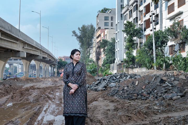 Bushra Afreen stands in mud and stones, rocks, and broken concrete beside an overpass and apartment buildings.