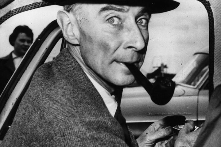 Robert Oppenheimer sits inside a car wearing a hat and smoking a pipe.
