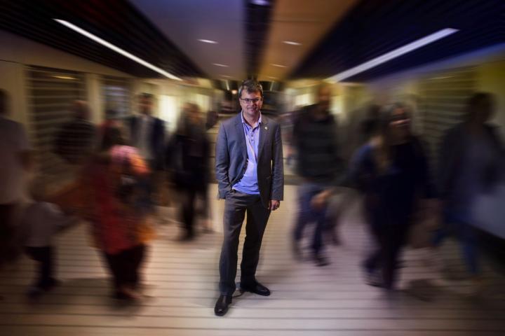 A full-body photo of Christian Leuprecht standing in a hallway facing the camera. The people to his left and right walking by are blurred.