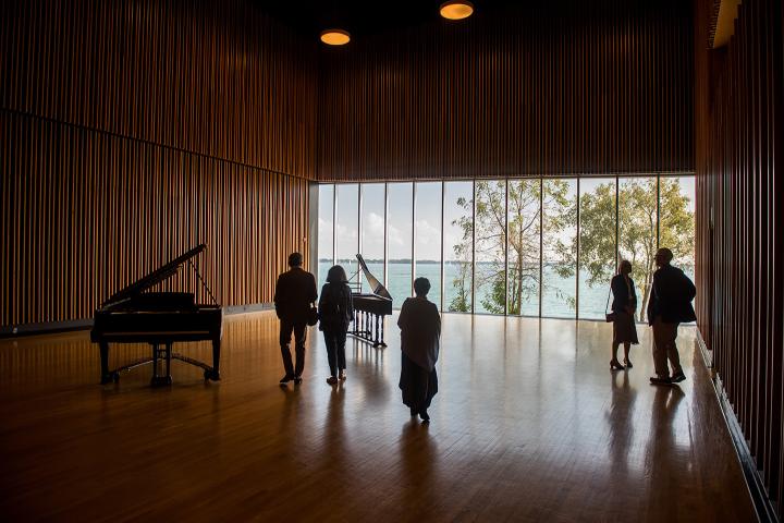 People wander around a room with wood floors, walls, and ceiling. Their are two pianos in the room and one wall of windows looks out on Lake Ontario.