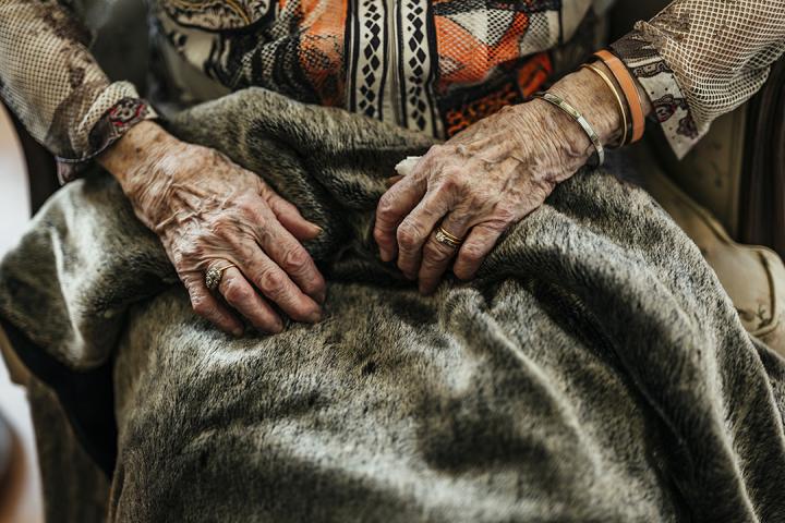 A close-up of an elderly woman’s hands resting on a soft fur blanket, showing her rings, bracelets, and patterned sleeves.