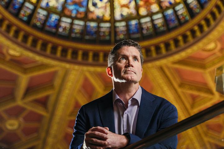 Portrait of a man in a suit standing beneath a stained-glass dome, looking upward with hands clasped on a railing.