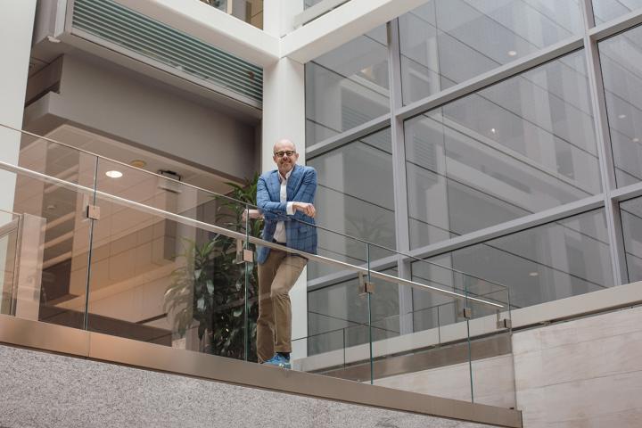 Man in a blue blazer stands on an upper-level balcony inside a modern glass atrium, leaning on the railing and looking down.