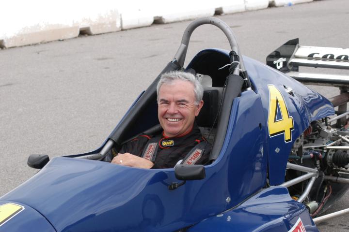 Man in a racing suit smiles while seated in a blue open-wheel race car marked with the number 4.