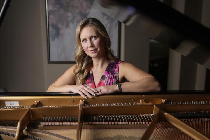 Woman leans on a grand piano, viewed across the open strings and soundboard, in a softly lit room.