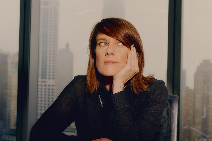Portrait of a woman seated at a table in front of large windows, resting her chin on her hand with a city skyline in the background.