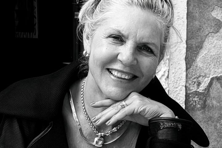 Black-and-white portrait of a smiling woman resting her chin on her hand, wearing a necklace and sunglasses perched on her head.