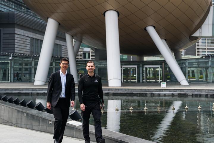 Two men walking beside a reflecting pool in front of a large futuristic oval-shaped building in an urban setting.