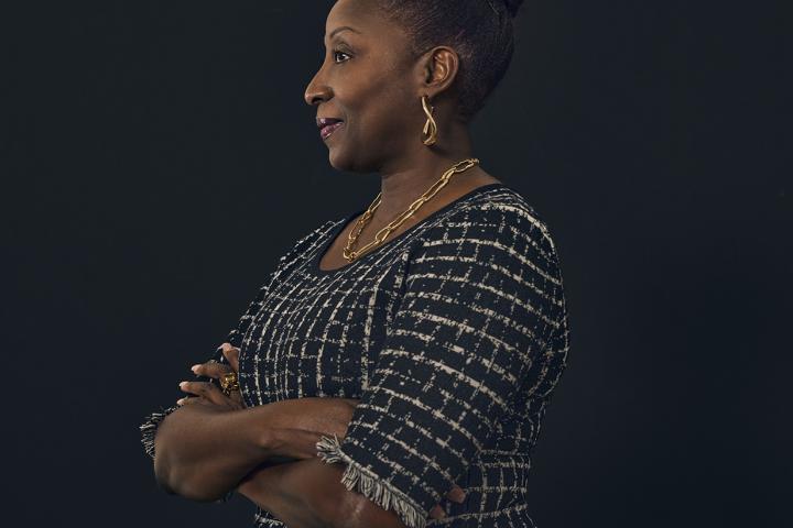 Profile portrait of a woman with arms crossed, wearing a patterned dress and gold jewelry against a dark background.