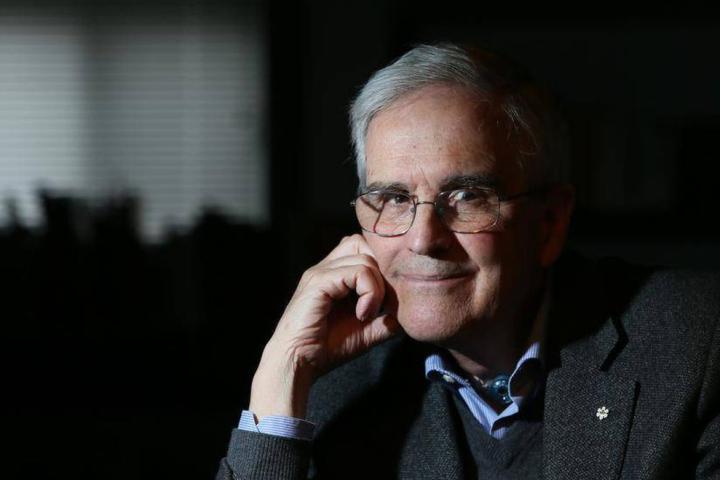 Portrait of an older man wearing glasses and a blazer, seated in low light with his hand resting against his face.