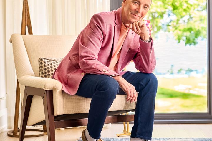 Man in a pink blazer seated in a bright room beside a large window.